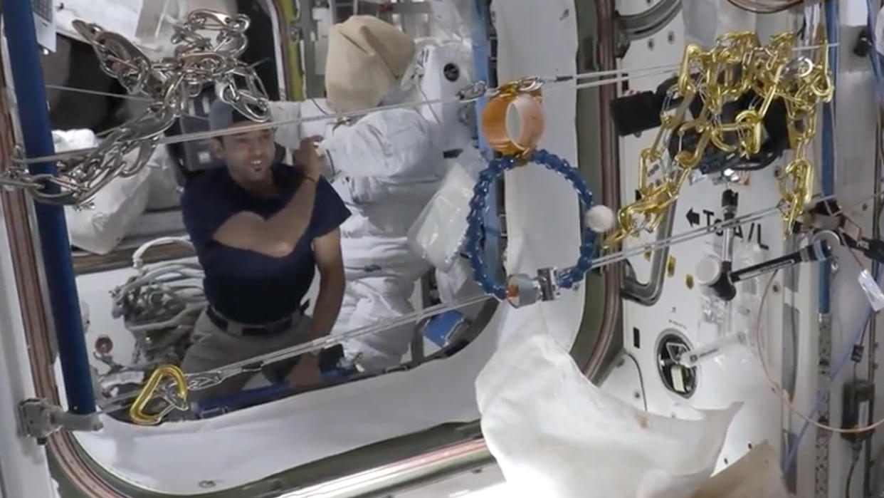  an astronaut floating in a space station module. he faces two tie down straps stretched across the entrance. between the two straps are two small rings, one above the other. he throws a ball through one of the rings. 