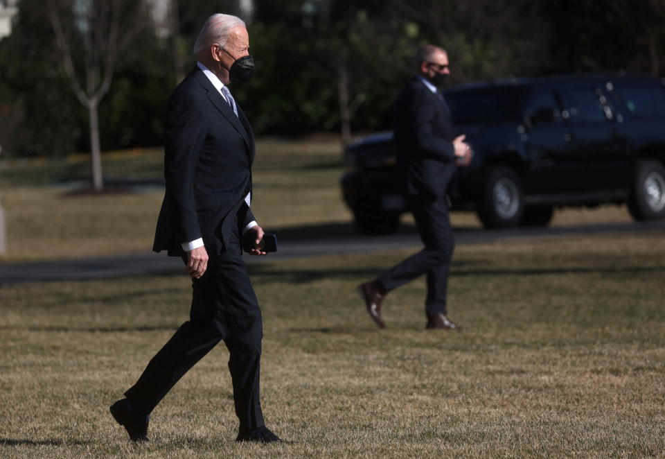 President Biden, wearing a mask, walks across the South Lawn of the White House. Another man, also masked, is seen in the background, yards away.