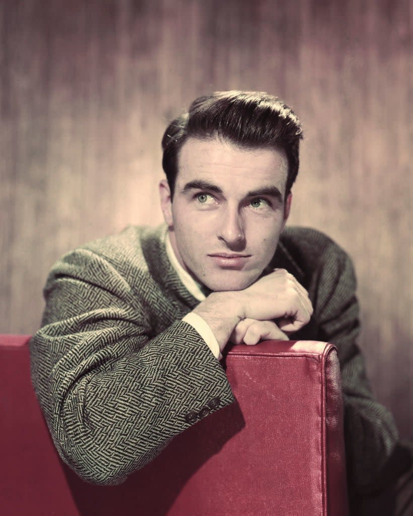 A man with neatly styled hair leans on a red cushion, looking thoughtful. He wears a patterned suit jacket and a collared shirt