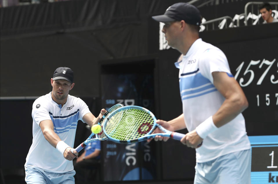 Mike Bryan, left, of the U.S. makes a backhand return as his brother Bob watches during their third round doubles match against Croatia's Ivan Dodig and Slovakia's Filip Polasek at the Australian Open tennis championship in Melbourne, Australia, Monday, Jan. 27, 2020. (AP Photo/Dita Alangkara)