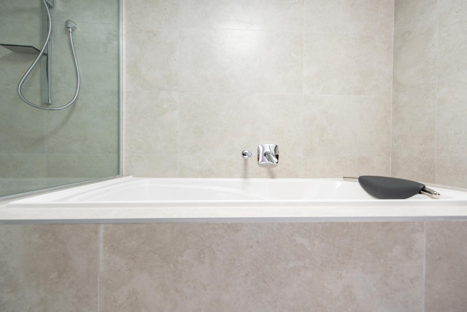 Modern bathroom with a spacious, empty white bathtub and a black bath pillow on its edge. A sleek showerhead is visible in the background