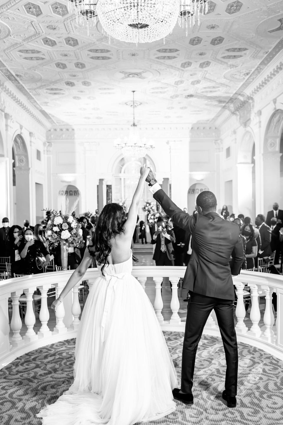 A black and white photo of a bride and groom raising their arms in celebration as they enter their wedding celebration.