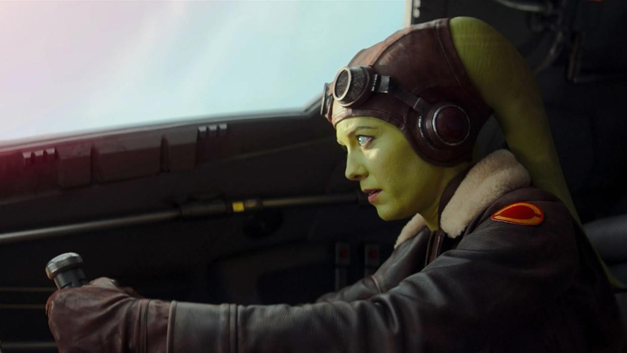  Hera Syndulla is a green-skinned Twi'lek with two head tails (known as lekku) protruding from the top left and right of her head. She is wearing a brown leather cap, goggles, and a brown leather aviator jacket with a fluffy collar. Here she is piloting a spaceship. 