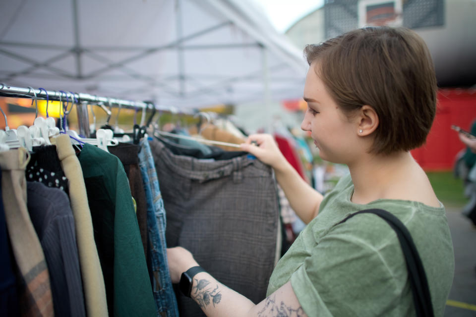 Person with short hair shopping for clothes at an outdoor market, holding a pair of trousers while looking at a rack of hanging garments