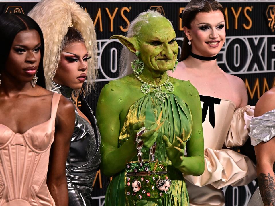 drag race queens on the emmys red carpet, with a person dressed as a goblin in a green dress, warts on her skin, her hair stringly, her face withered