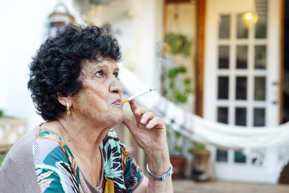 An elderly woman with curly hair is sitting outside, smoking a cigarette while looking into the distance. She wears a patterned blouse and gold earrings