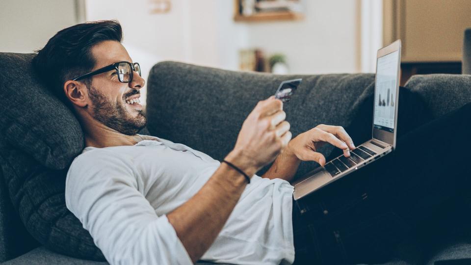 Smiling man lying on the couch and shopping online with credit card and laptop.