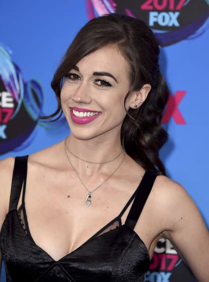 Colleen Ballinger with bangs and a ponytail wearing a scrappy black dress against a blue backdrop