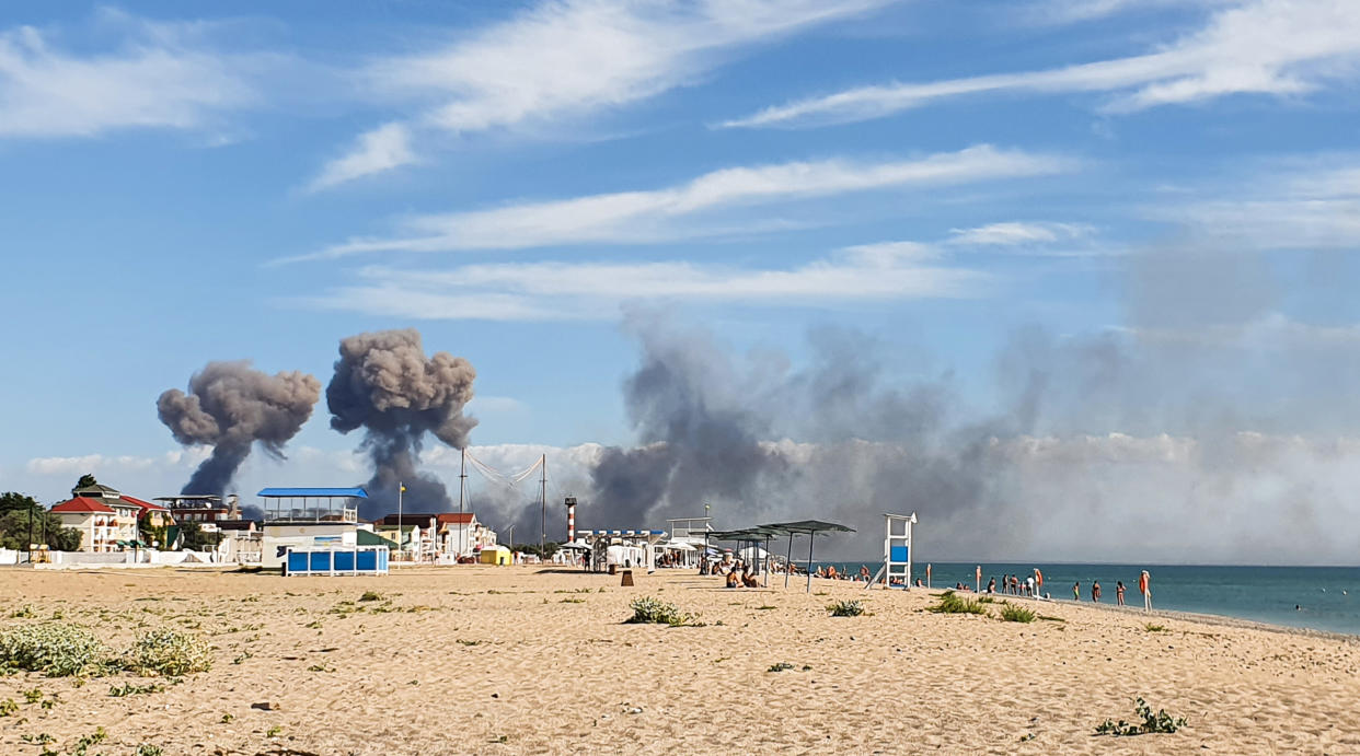 Plumes of smoke rise in the distance in a photo taken from a beach.