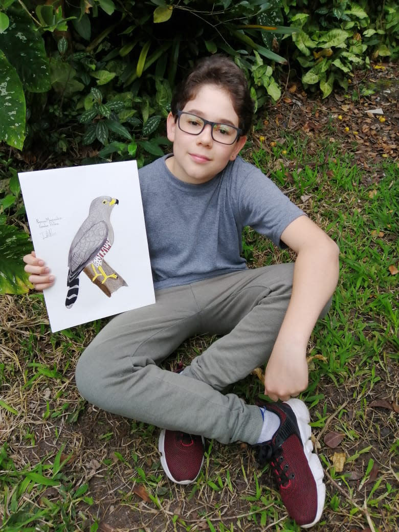 In this Aug. 2020 photo provided by Johana Reyes Herrera, Jacobo Rendon, 14, poses with his illustration of an Roadside Hawk in his backyard in El Camino de Viboral, Colombia. Rendon has been working on a photographic and illustrated bird guide that he plans to donate to a local cultural center. (Johana Reyes Herrera via AP)