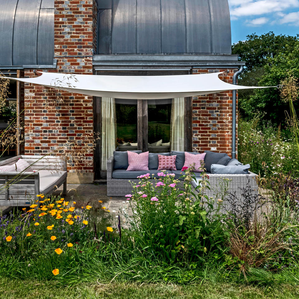 White sail shade above a flagstone patio area next to a red brick house with a wicker corner sofa lined with grey and red cushions