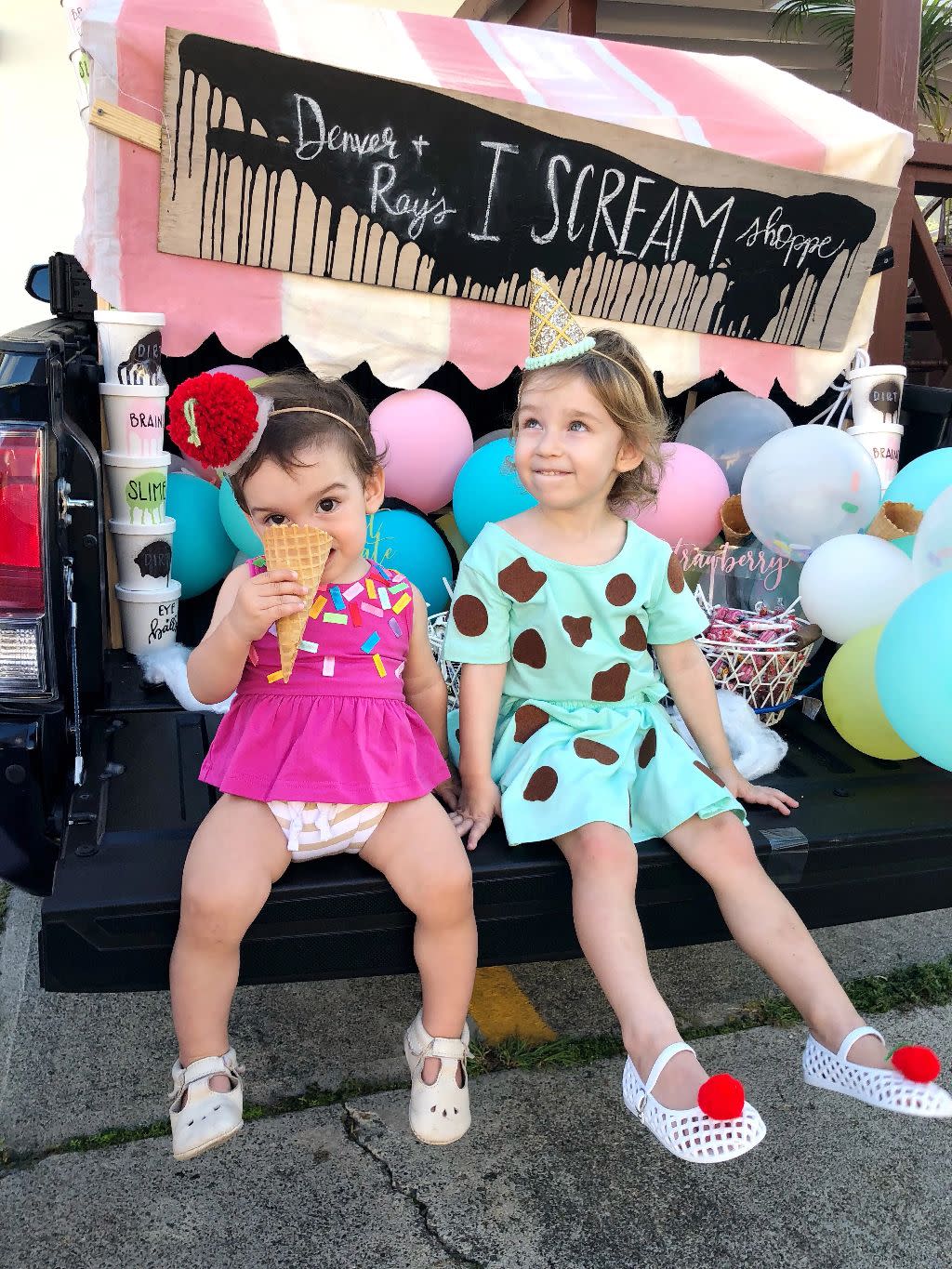 two little girls sitting on tailgate of truck one is wearing a pink dress with colorful sprinkles and eating a waffle cone the other is wearing a mint green dress with brown shapes like chips on it there are pastel colored balloons in the bed, a pink and white awning over it with a banner than reads i scream for ice cream and ice cream pint containers with labels like slime stacked on one another