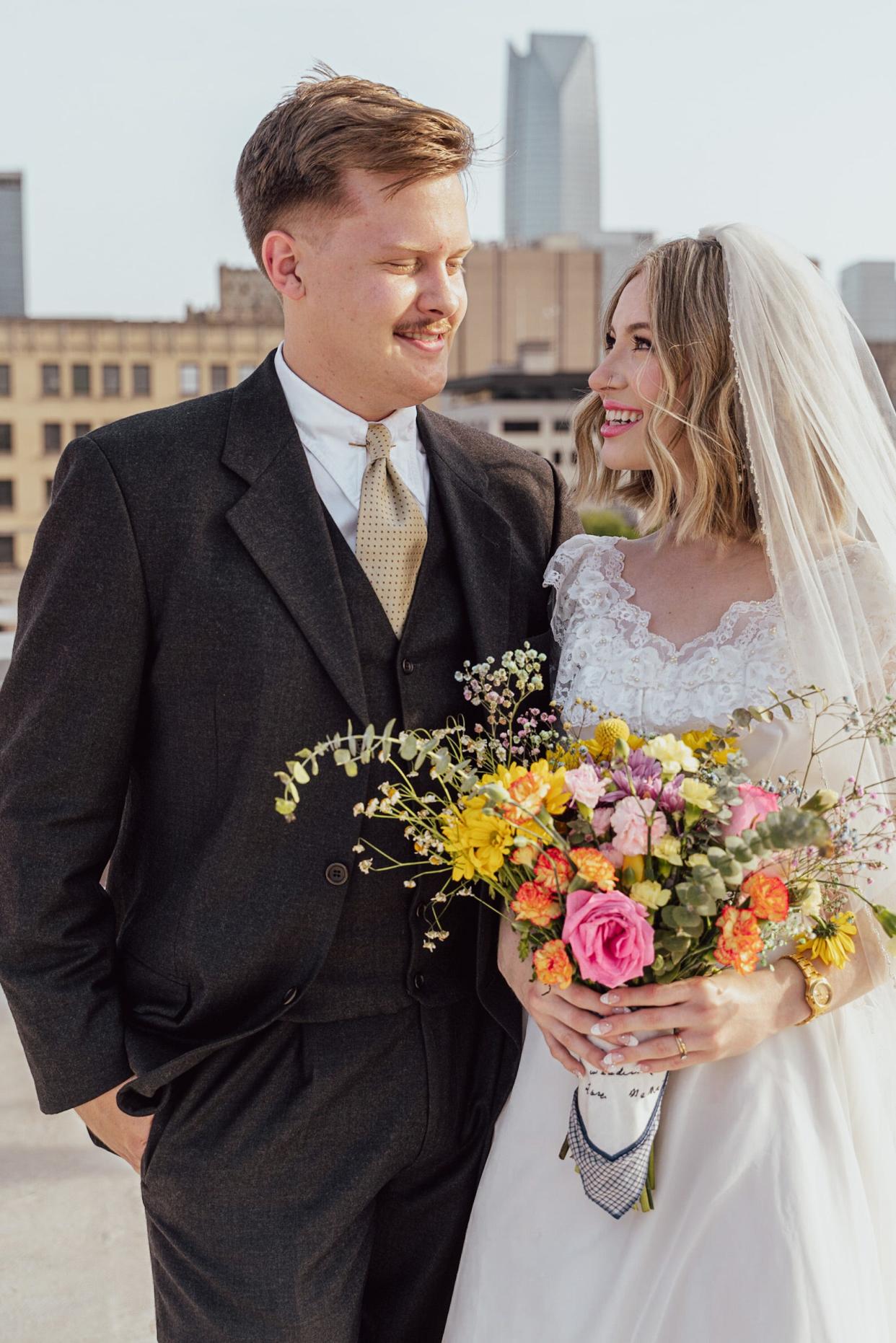 A bride and groom smile at each other on a rooftop on their wedding day.