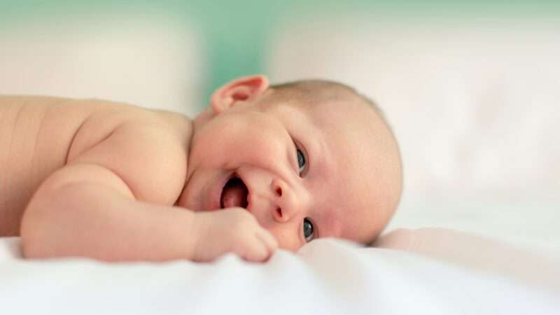 A baby smiles while lying on a white blanket on a bed with a green wall in the background