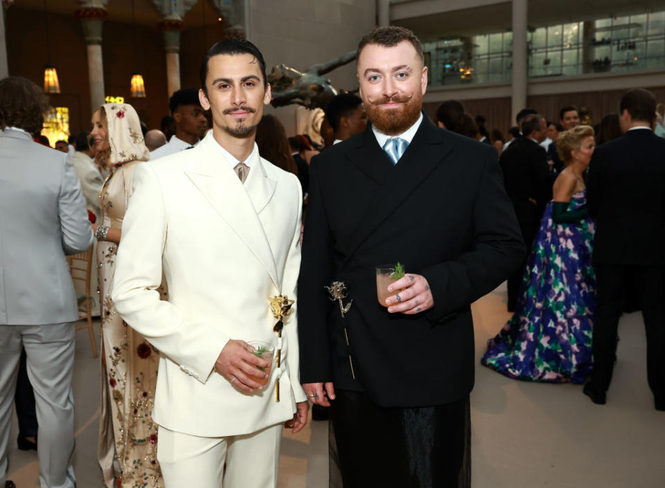 Two men at an event, one in a white suit holding a trophy, other in a black suit with a drink, standing indoors