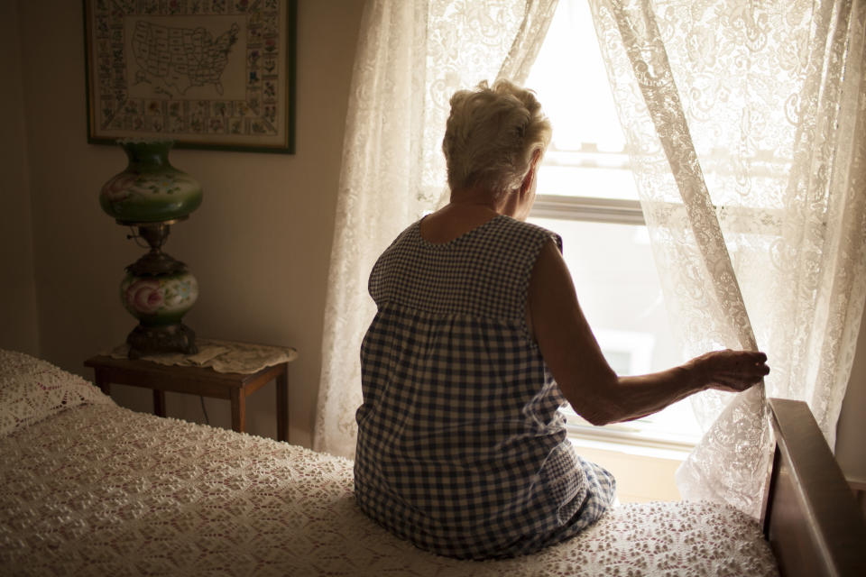 Old woman sits on bed while looking out the window. 