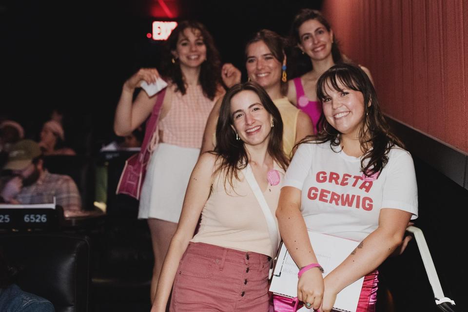 a group of women standing in the aisle of a movie theater, smiling and wearing pink. One of the women in front wears a t-shirt that says "Greta Gerwig."