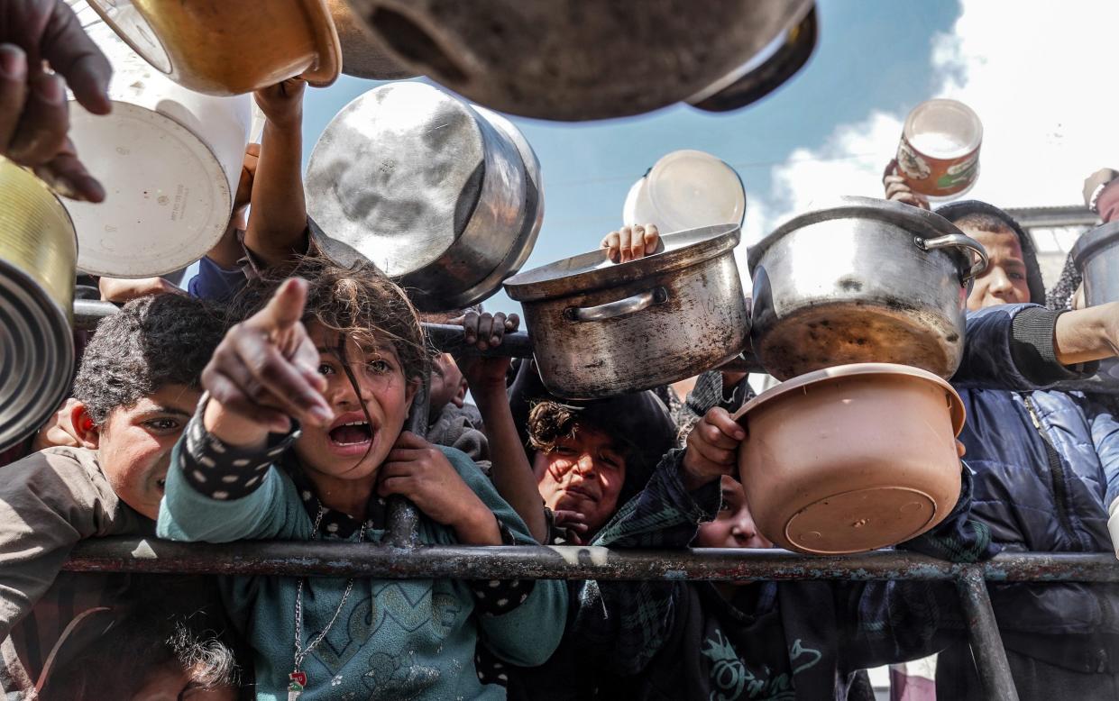 Palestinian children, holding empty pots, wait in line to receive food prepared by volunteers