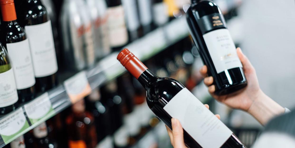 over the shoulder view of woman walking through liquor aisle and choosing bottles of red wine from the shelf in a supermarket