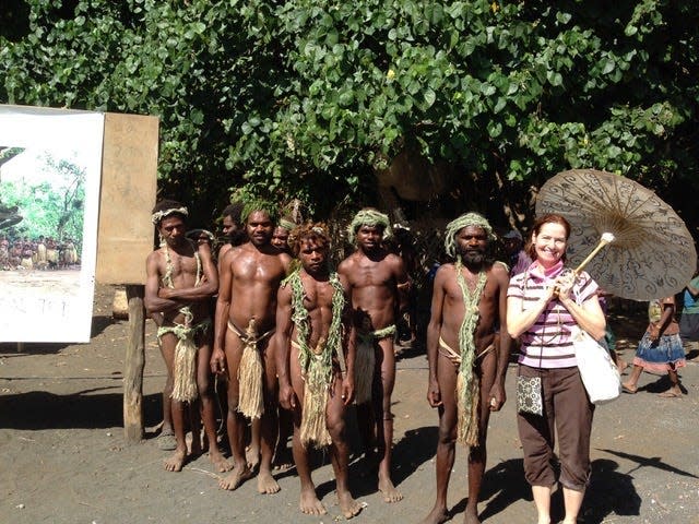 Whitmire, holding an umbrella, poses outdoors in front of a group of men wearing loincloths made of leaves. Some have headpieces and necklaces also made of leaves.