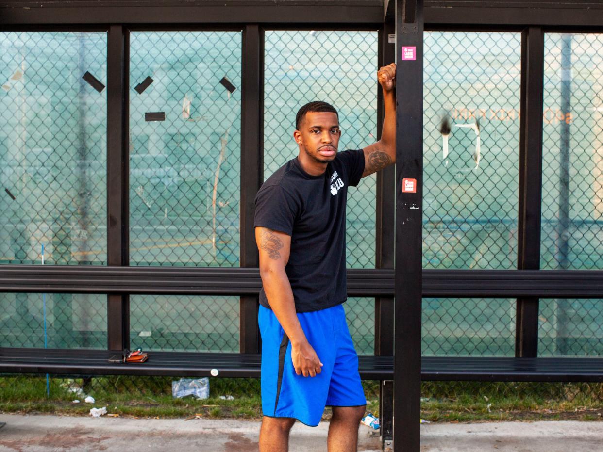 Amazon Labor Union vice president Derrick Palmer stands at a bus stop in front of a Staten Island Amazon warehouse he helped unionize. He is wearing a black t-shirt and blue basketball shorts. Stickers supporting the ALU are on the bus stop.
