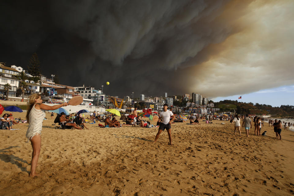 Turistas juegan a las palas en una playa con una gran nube de humo negro a sus espaldas, procedente de los incendios forestales, en Viña del Mar, Chile, el viernes 2 de febrero de 2024. (Martín Thomas, Aton Chile vía AP)