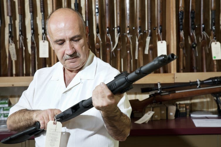 A man cocks a rifle, standing in front of a rack of rifles in a US gun shop.