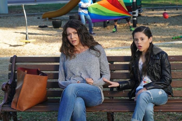 Two people are sitting on a wooden park bench. One is wearing a gray sweater; the other, a black leather jacket. A rainbow-colored kite is flying in the background