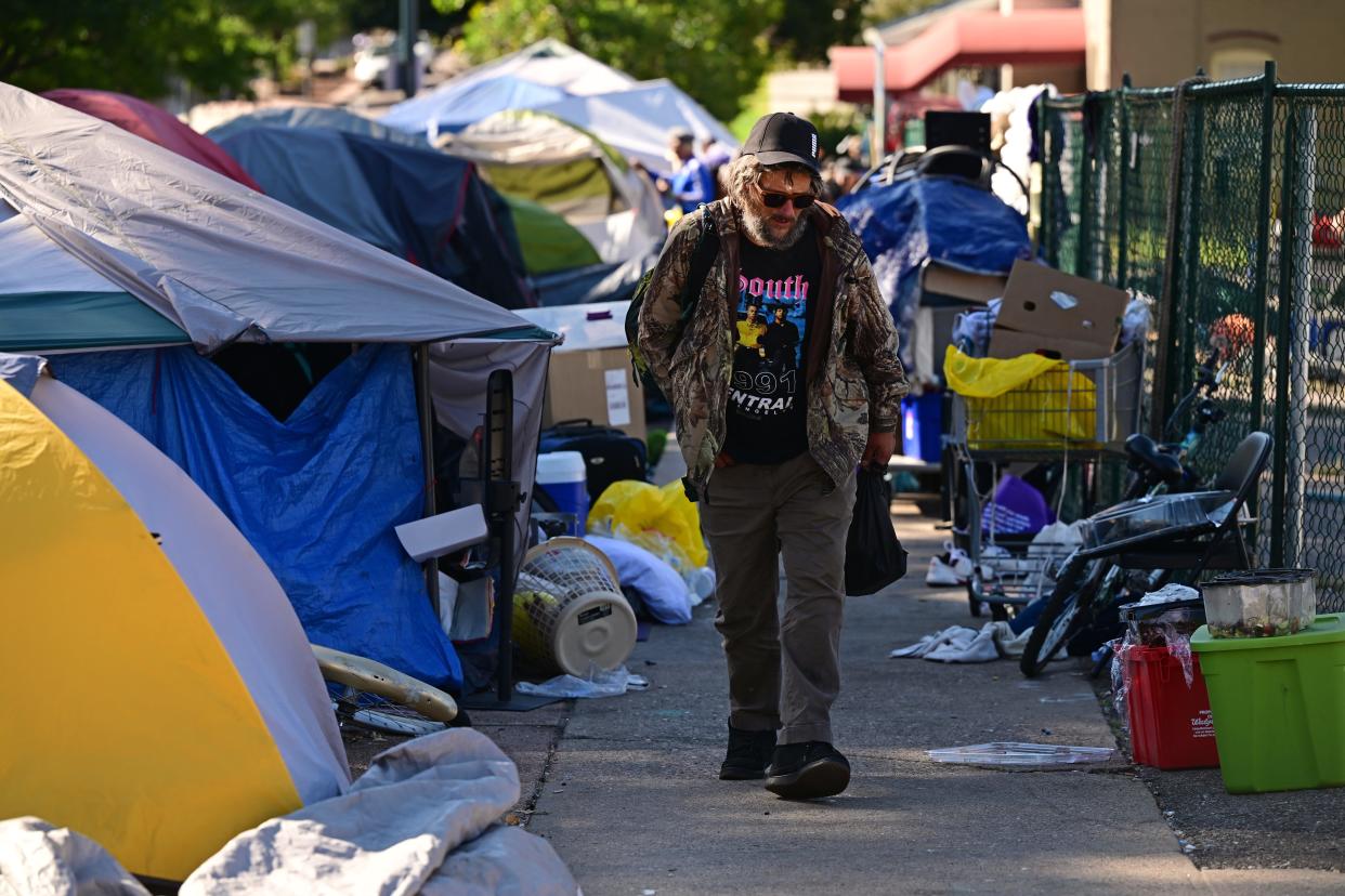 A bearded man wearing a graphic tee and sunglasses walks toward the camera with tents and debris to his left and right