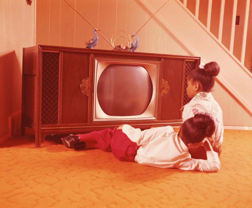 Two children with updos are watching a vintage TV in a retro living room with orange carpet and wood paneling, evoking a nostalgic setting