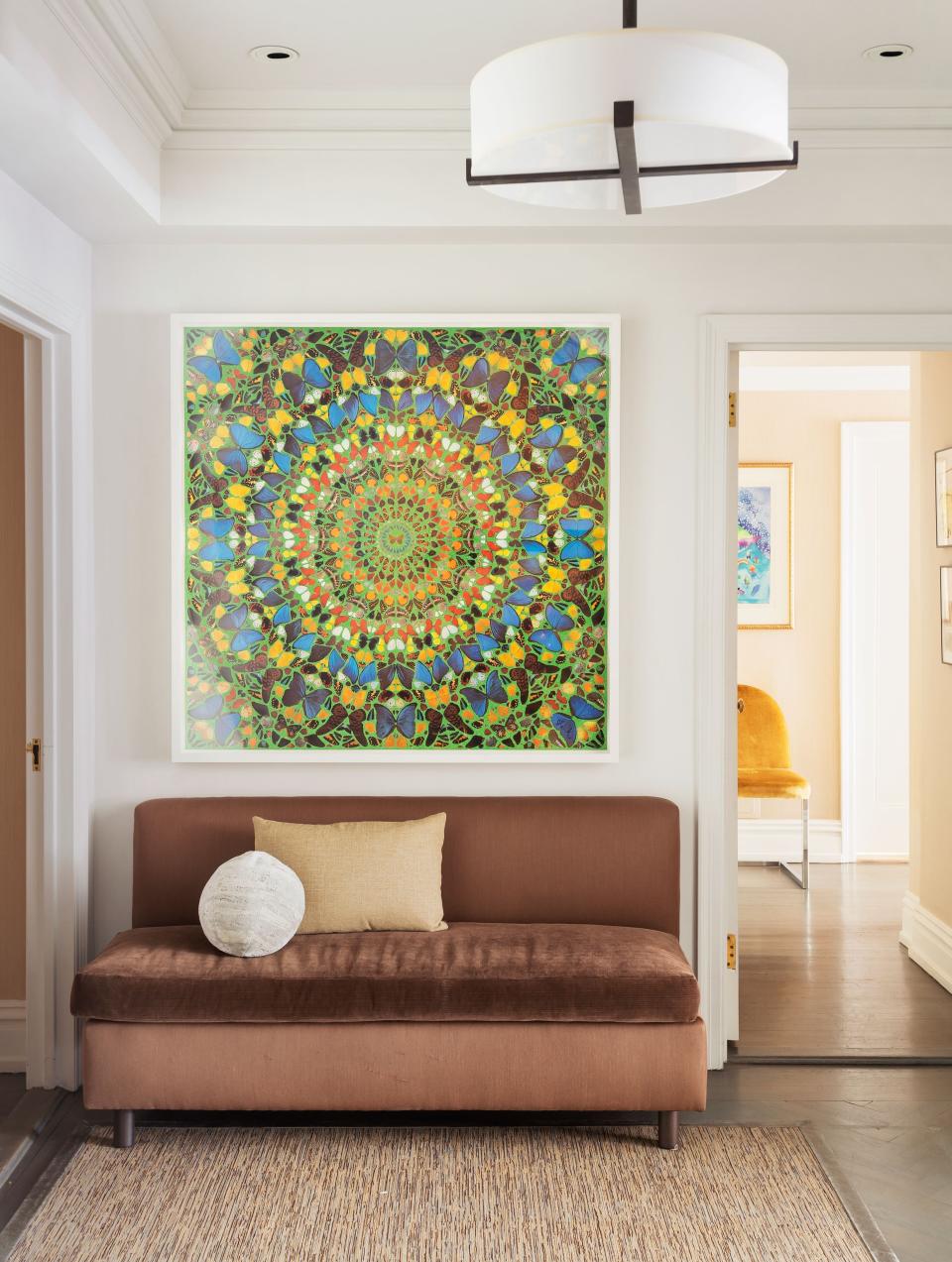 The apartment’s entryway features a custom armless banquette upholstered in Holland & Sherry velvet, and a custom metal and parchment pendant designed by Huniford. The striking artwork is by Damien Hirst.