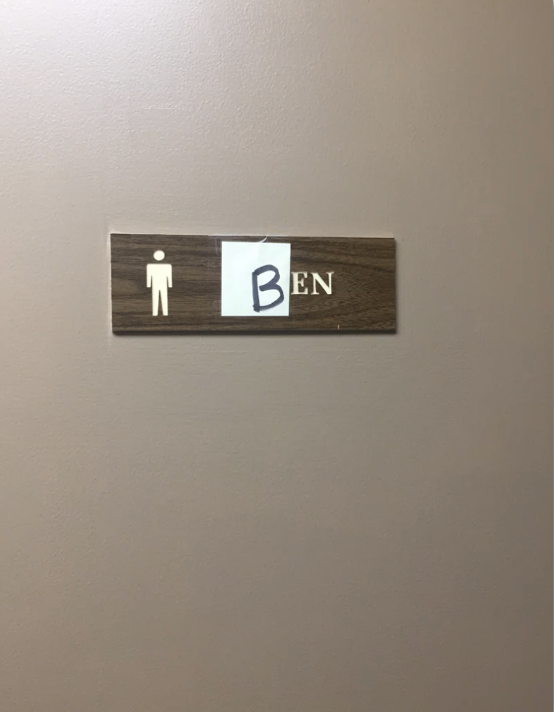 A restroom sign, with a handwritten paper note covering part of the name, forming "Ben" next to a men's restroom symbol