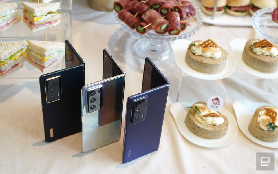 <p>The black, silver and purple versions of Honor Magic V2 surrounded by sandwiches, cakes and other finger foods.</p>
