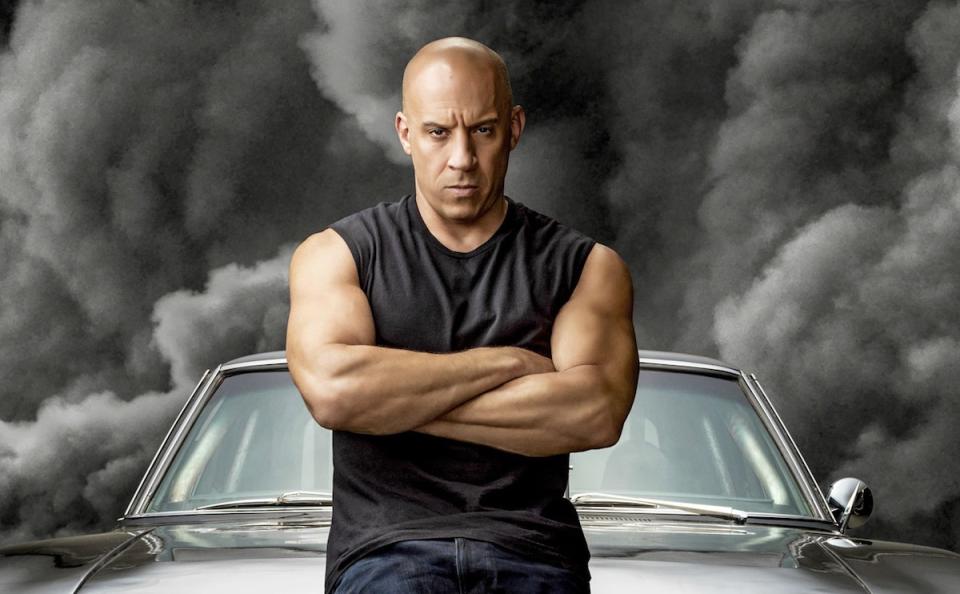 Vin Diesel in a black sleeveless t-shirt standing against a black car with black smoke behind it