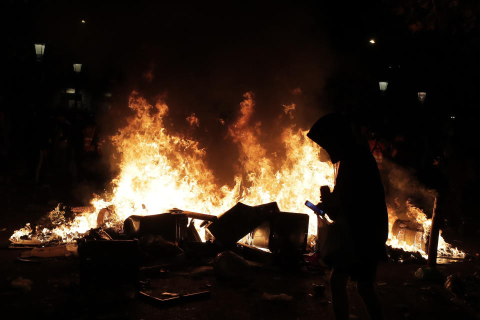 A protester looks at a cell phone by a burning trash container that was set on fire in Barcelona, Spain, Thursday, Oct. 17, 2019. Catalonia's separatist leader vowed Thursday to hold a new vote to secede from Spain in less than two years as the embattled northeastern region grapples with a wave of violence that has tarnished a movement proud of its peaceful activism. (AP Photo/Manu Fernandez)
