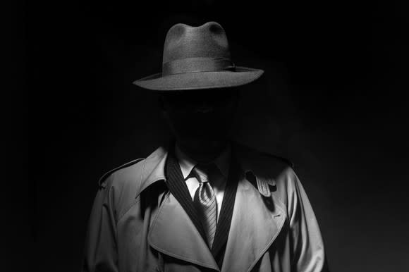 A person whose face is obscured wearing a trenchcoat, suit, tie, and fedora.