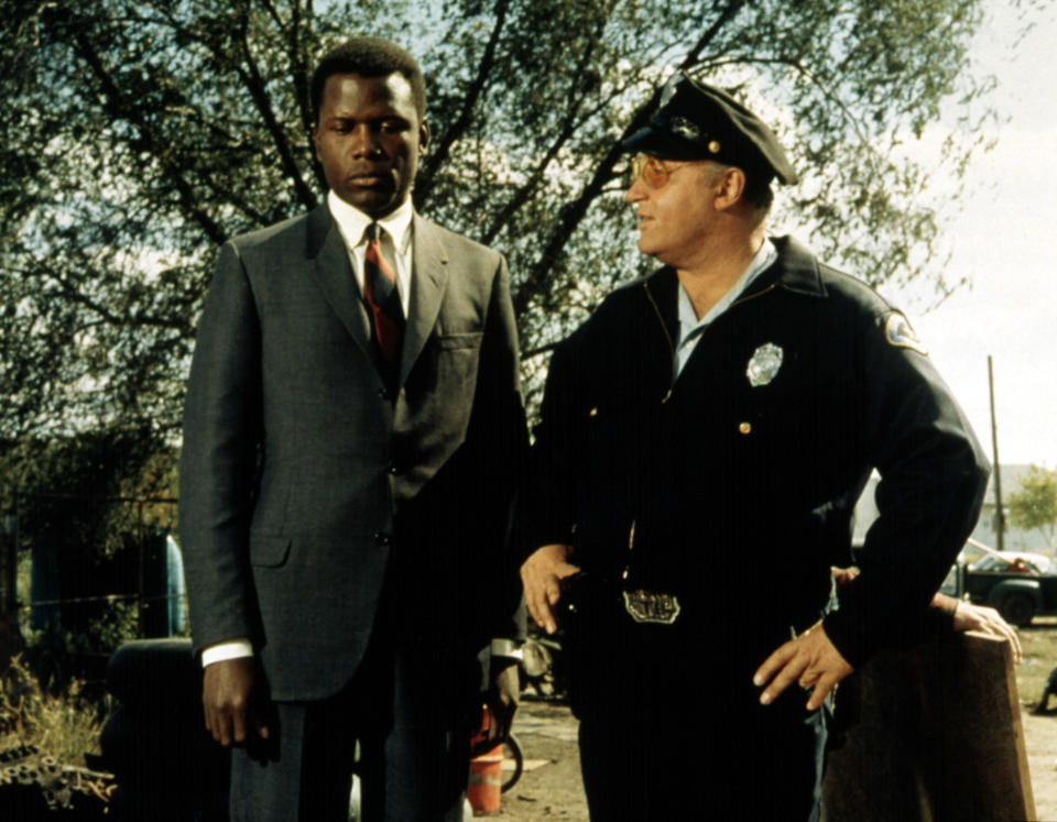 Sidney Poitier, in a grey suit, stands next to Rod Steiger, dressed as a police officer, in an outdoor setting with trees in the background