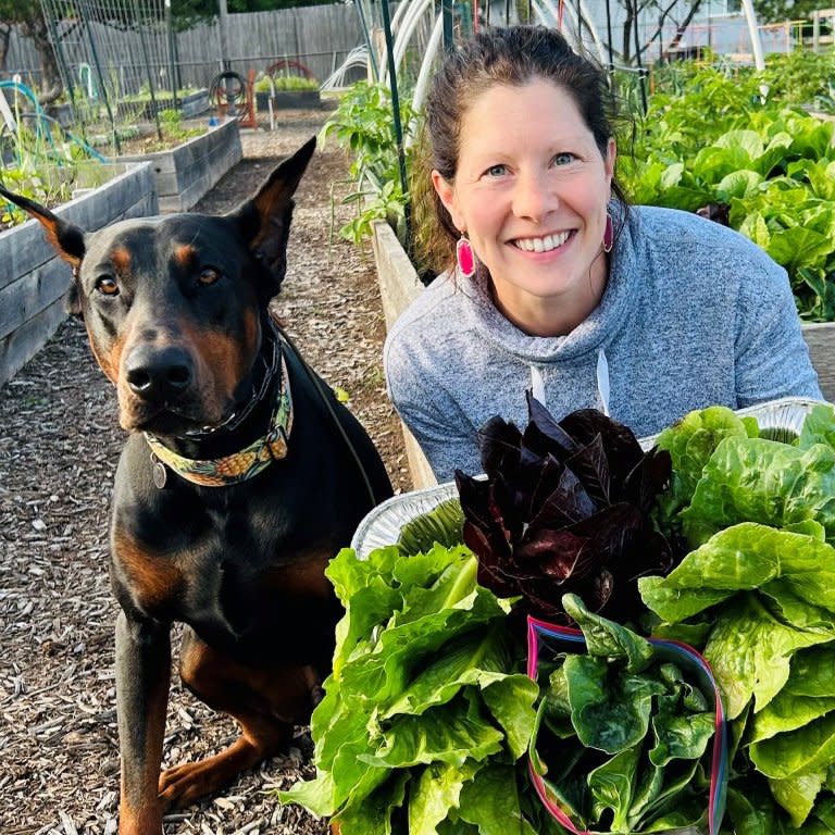  A smiling woman with her hands full of lettuce next to a dog. 