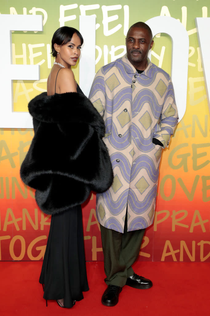 Two people pose on the red carpet; one wearing a black gown with a large fur shawl, the other in a patterned jacket and trousers