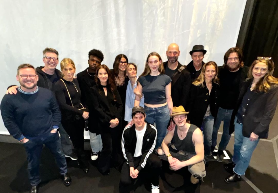 Group photo of multiple people smiling, casual attire, some seated, others standing, in an indoor setting