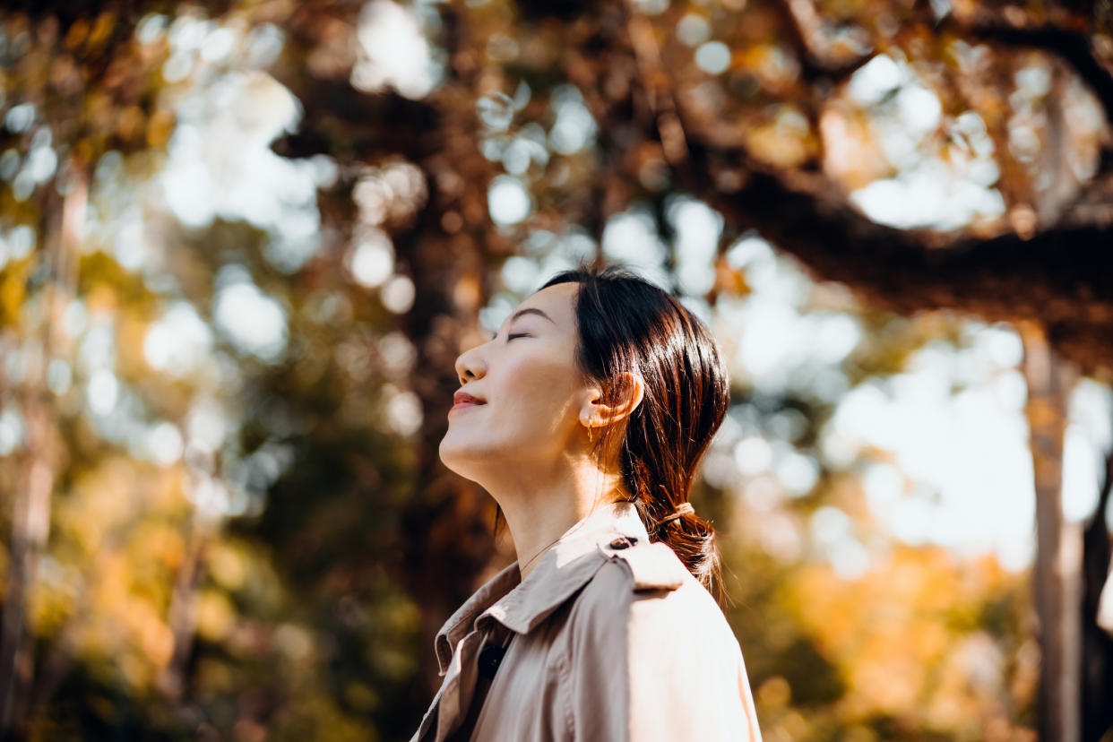 Portrait of young Asian woman having a walk in the park, enjoying the warmth of sunlight on a beautiful Autumn day outdoors and breathing fresh air with eyes closed. Relaxing in the nature under maple trees