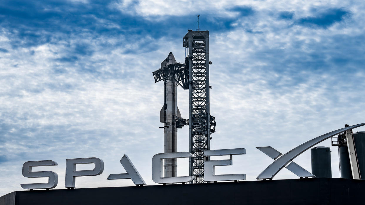  A large silver rocket stands next to its launch tower, with a large sign reading "spacex" in the foreground. 