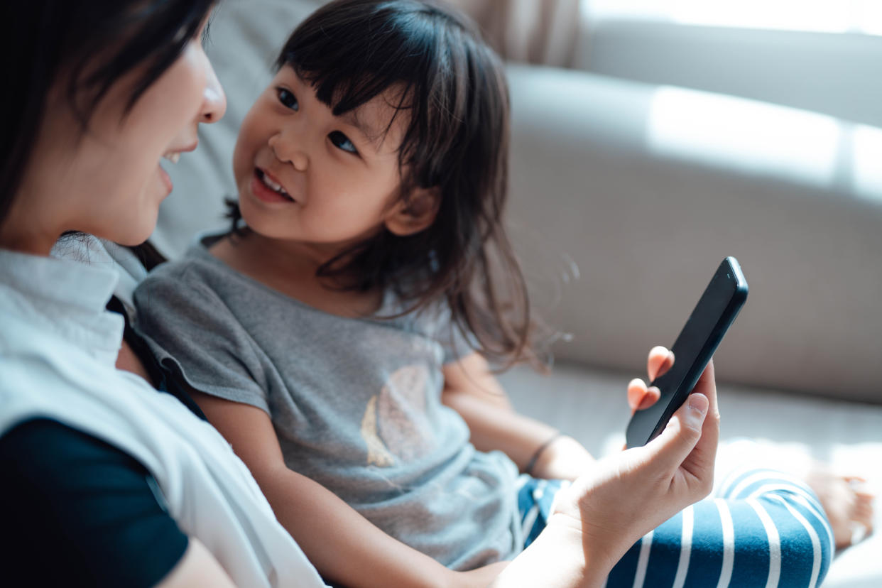 Cropped shot of young Asian mom and daughter having video call on cell phone with their family, sitting on sofa together during the day. Family Time with Technology.