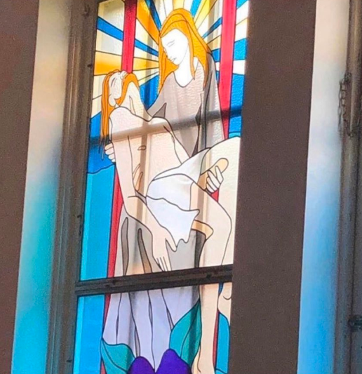 A photo of a stained-glass church window with Virgin Mary holding Jesus by a peculiar phallic object.