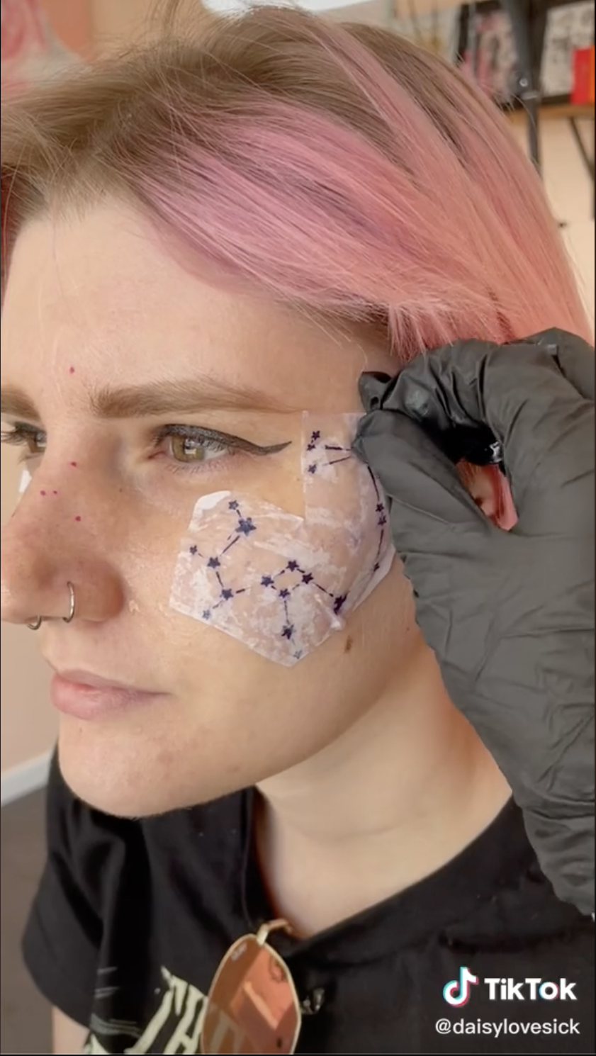 Screengrab from a TikTok by user daisylovesick of a hand placing a tattoo stencil of a constellation on a client's face