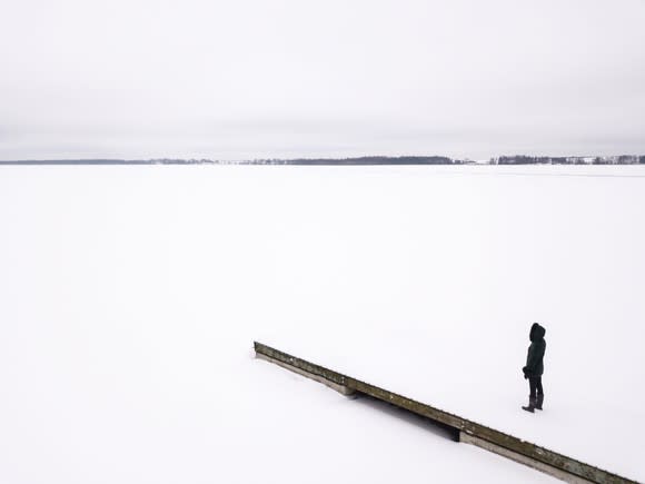 Woman in winter outerwear standing on snow-covered pier overlooking a frozen lake.