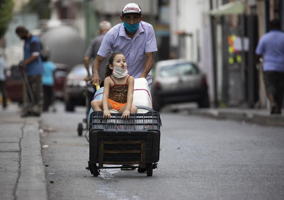 Un hombre, con mascarilla para protegerse del coronavirus, empuja un carrito lleno de recipientes de agua vacíos mientras va, acompañado de una niña, en busca de agua, en Caracas, Venezuela, el 20 de junio de 2020. Sin agua corriente en sus casas, los residentes no tienen más opción que salir a buscarla, violando la cuarentena decretada en todo el país para combatir la pandemia. (AP Foto/Ariana Cubillos)