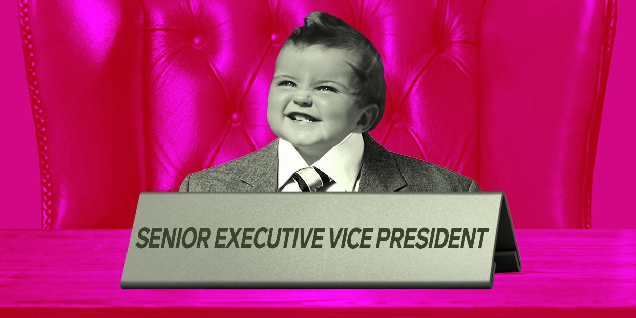 A baby wearing a too-large suit sits at a desk with a job-title plaque in front of them that reads 'Senior Executive Vice President"