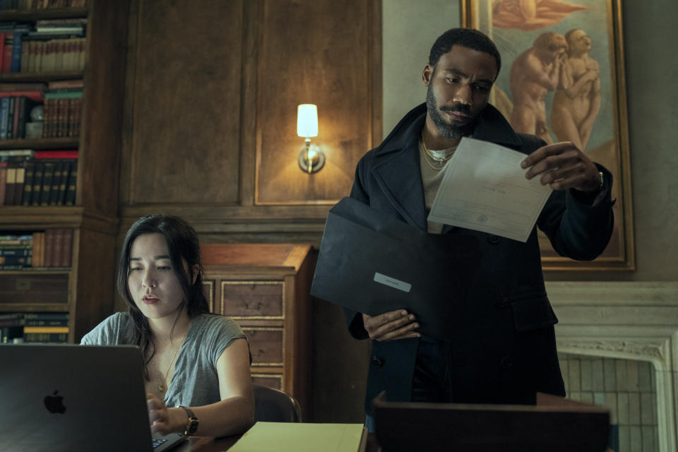 Maya Erskine sentada em frente a um laptop.  Donald Glover olha um documento.