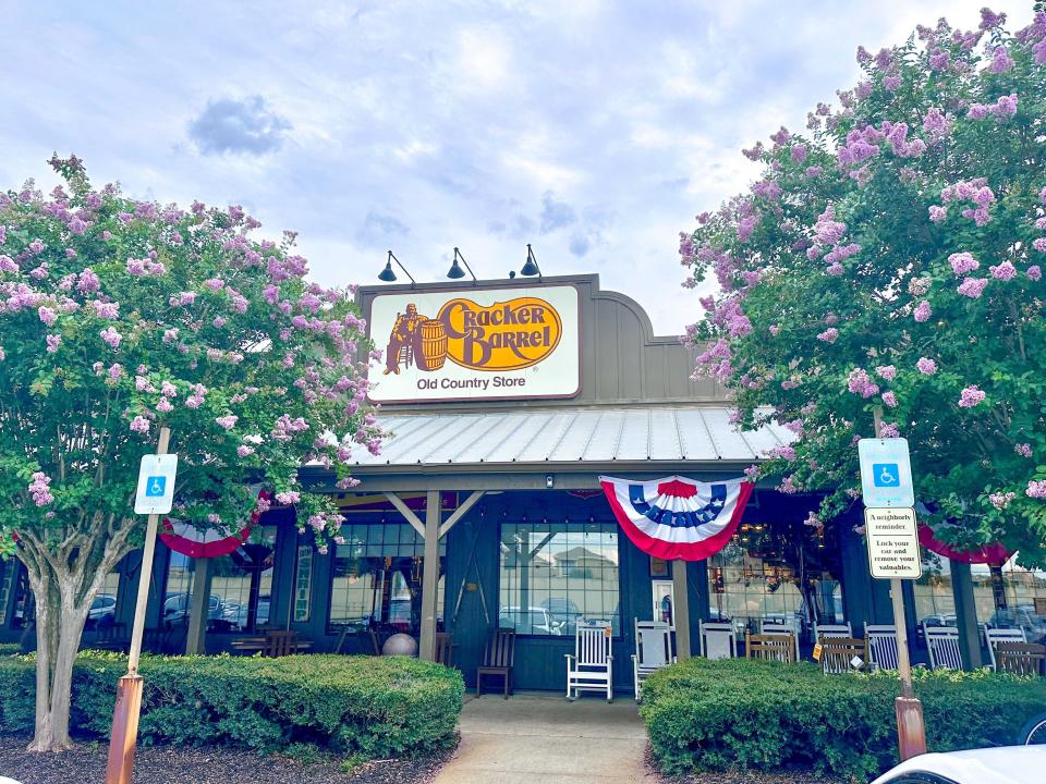 Exterior of Cracker Barrel with several trees with purple flowers surrounding the building. A sky filled with clouds is in the background.